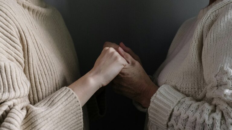 Close-up of an adult daughter and older mother holding hands, wearing cozy knit sweaters in warm neutral tones.