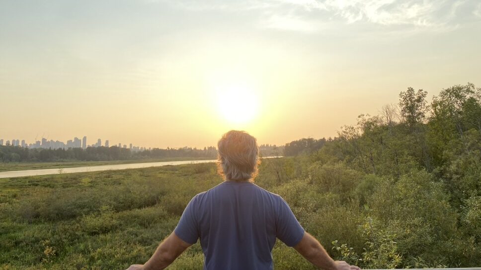 Man standing at a railing overlooking a river and city skyline at sunset, reflecting quietly — symbolizing nervous system regulation and shifting from self-criticism to self-respect.